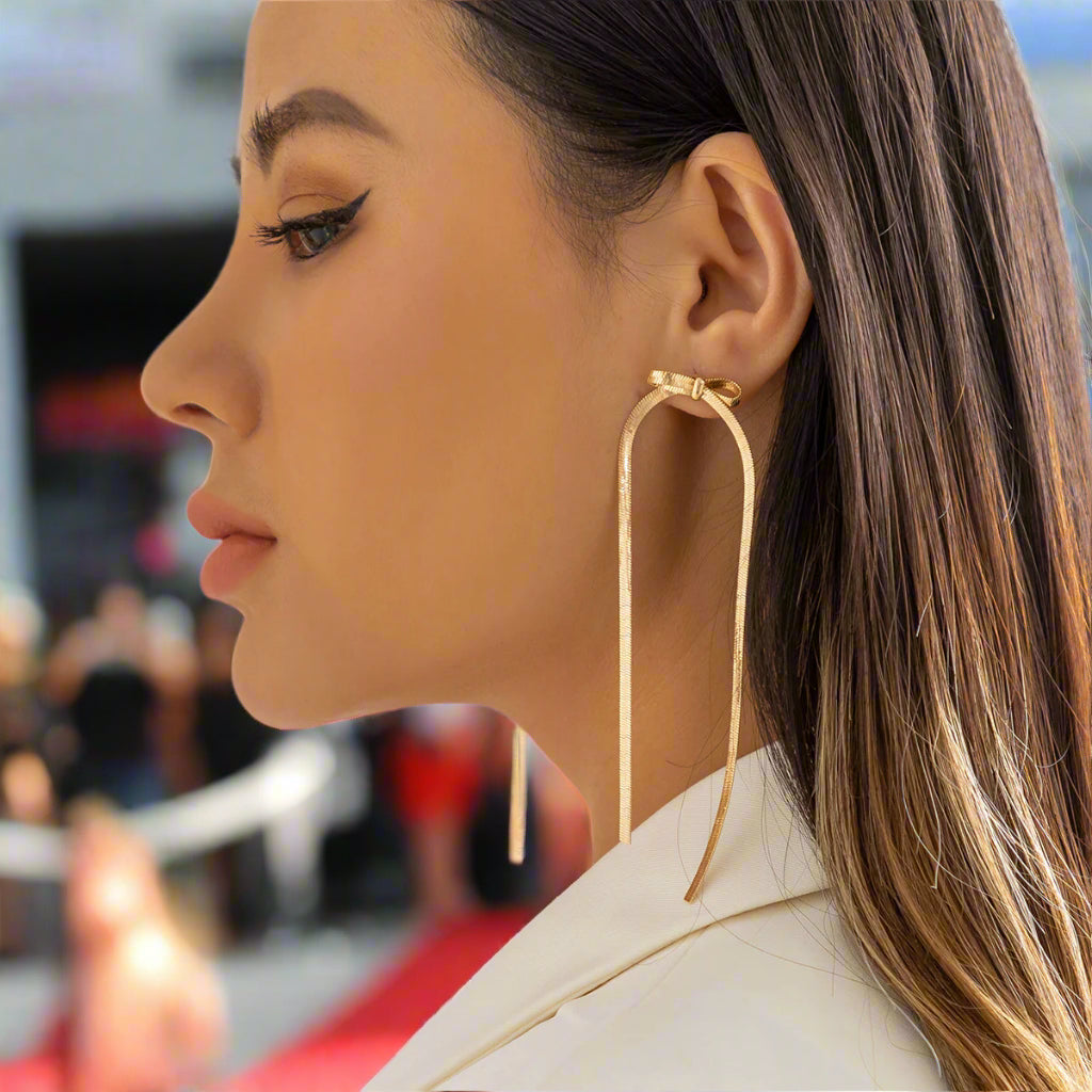 A womanwearing gold-colored herringbone chain dangle earrings with a bow design.