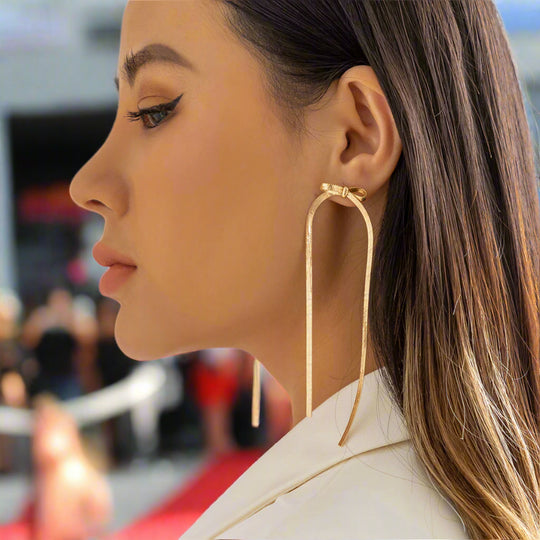 A womanwearing gold-colored herringbone chain dangle earrings with a bow design.