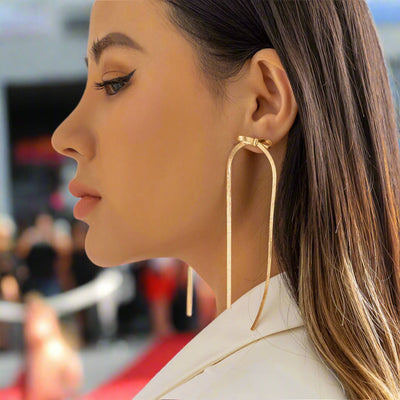 A womanwearing gold-colored herringbone chain dangle earrings with a bow design.