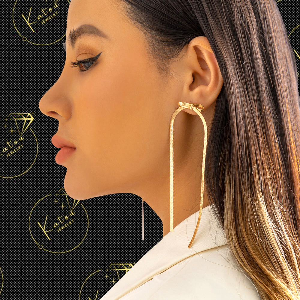 A woman wearing gold-plated bow herringbone chain dangle earrings on a black background with the Katou Jewelry logo patterns.